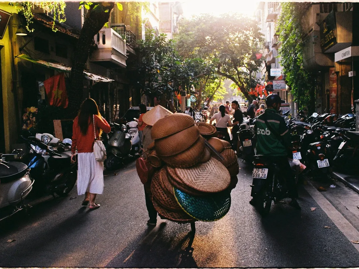 A busy street with people walking and riding motorcycles, and a man with a cart full of luggage.