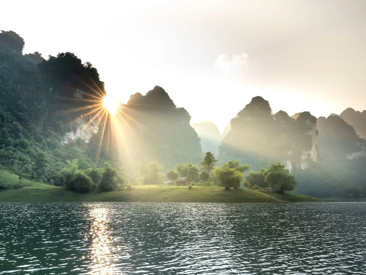 A serene scene of a lake with a sun shining on the water and a mountain in the background.