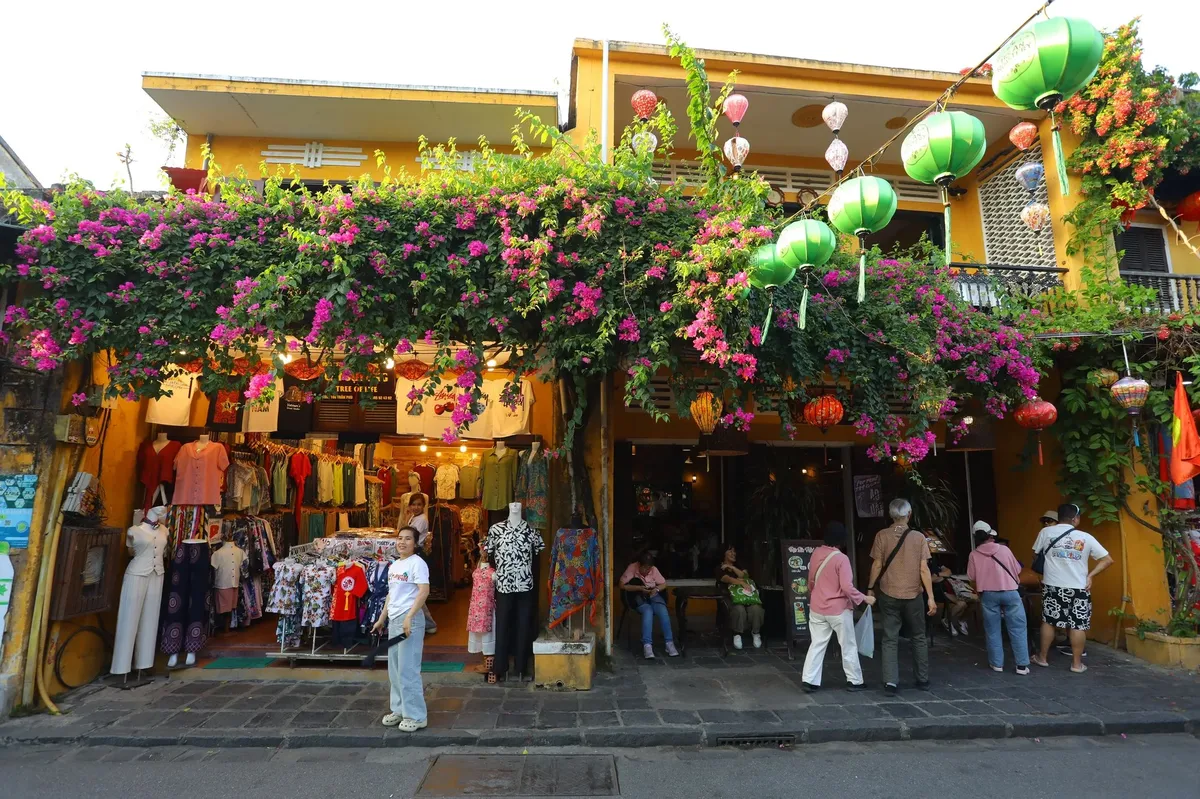 A busy street with a store front full of colorful clothing and a large tree with pink flowers hanging over it.