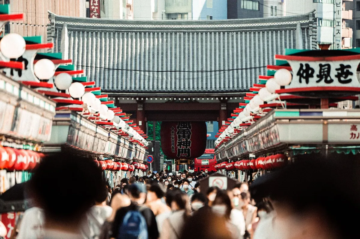 A busy shopping area with many people walking under a bridge.