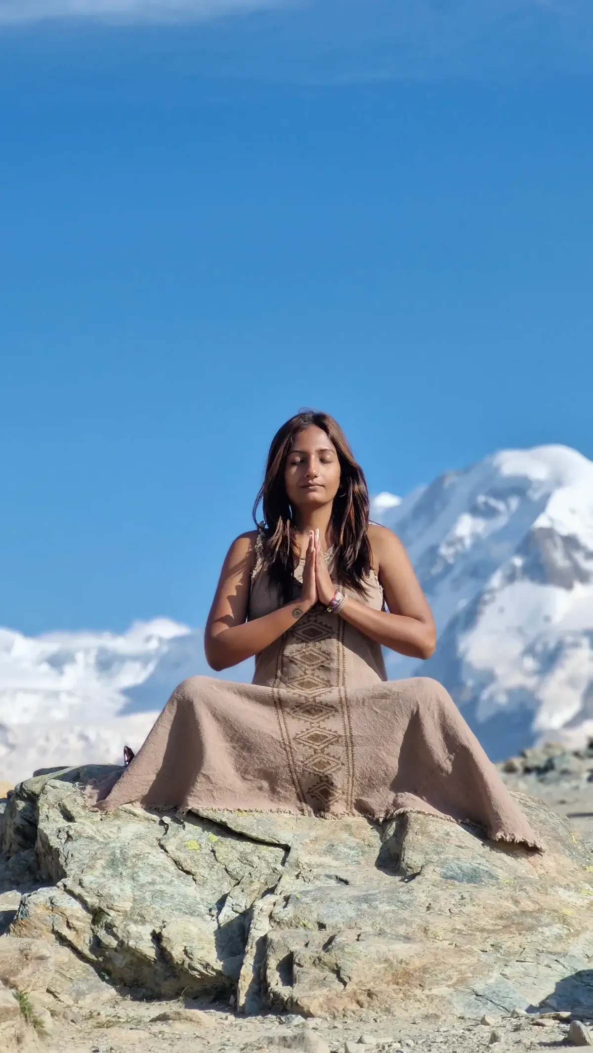 A woman in a brown dress is sitting on a rock, praying in front of a mountain.